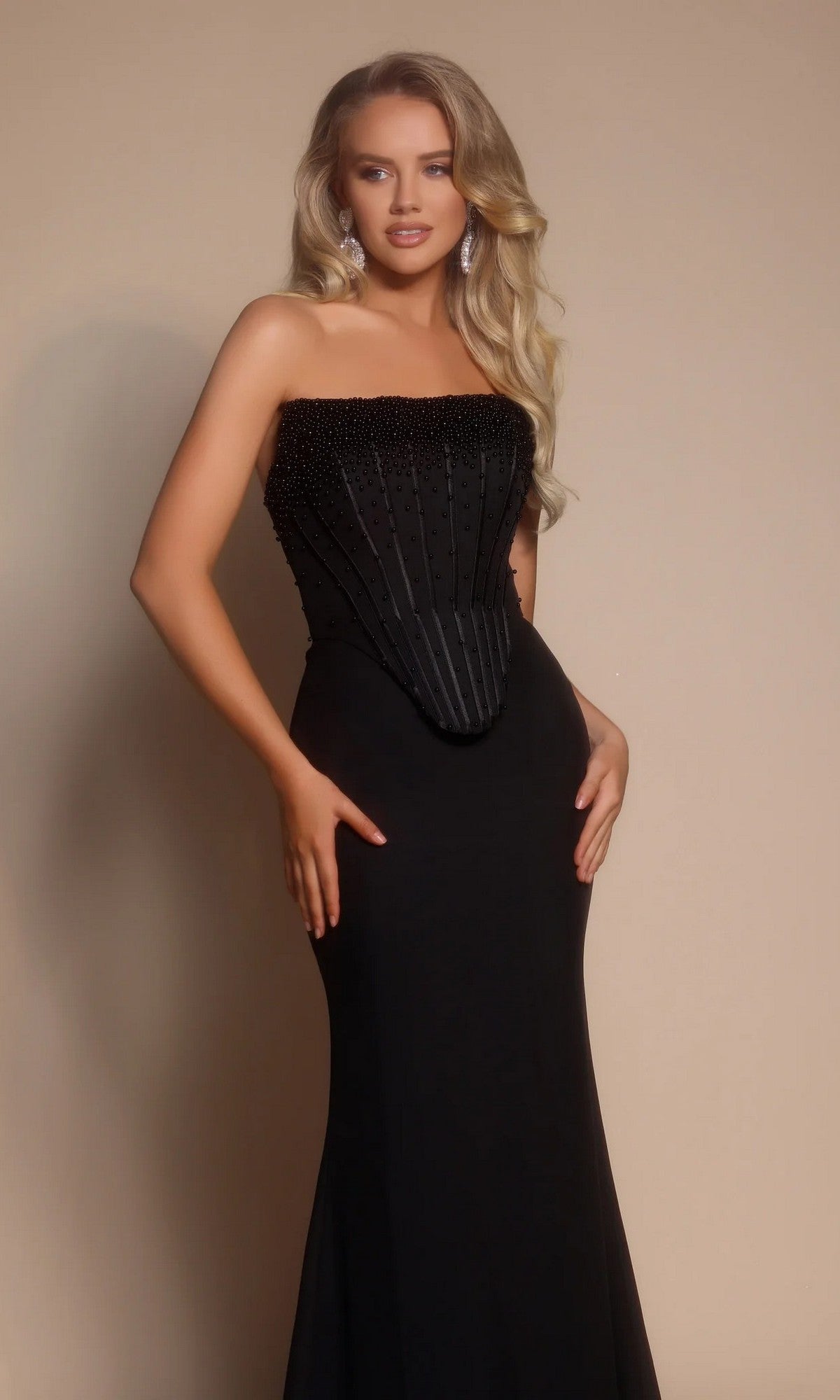 A woman models the Portia and Scarlett PS26008 long formal dress, featuring a strapless, fitted black silhouette with a beaded corset bodice and floor-length skirt, against a plain beige background.